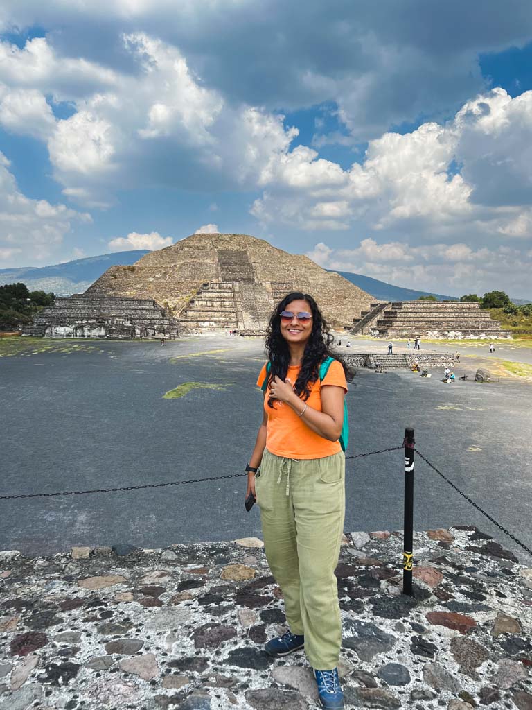 A woman wearing orange t-shirt and green pants, posing at Teotihuacan archaeological site, with the Pyramid of the Moon in the background.