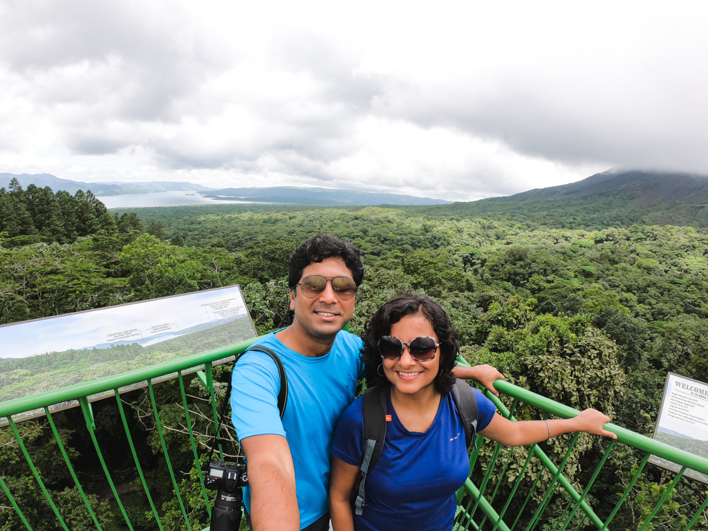 A man and woman standing on the observation tower with the rainforests of Arenal and Arenal Lake in the background