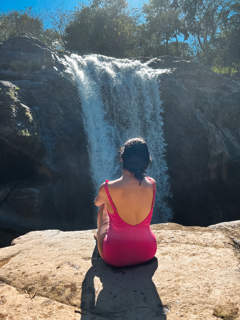 A woman in pink swimsuit, sitting on the edge of the rock, admiring the view of the waterfalls before her.