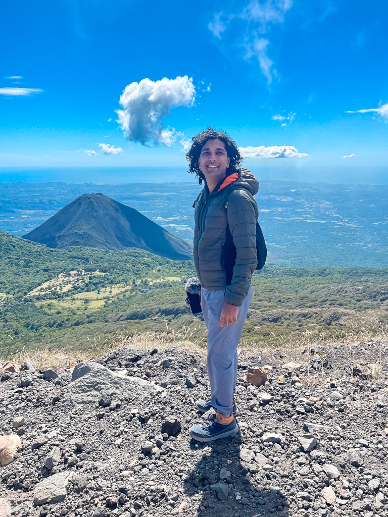 A man wearing a jacket and smiling at the camera, at the summit of the Santa Ana volcano. Izalco volcano seen in the background.
