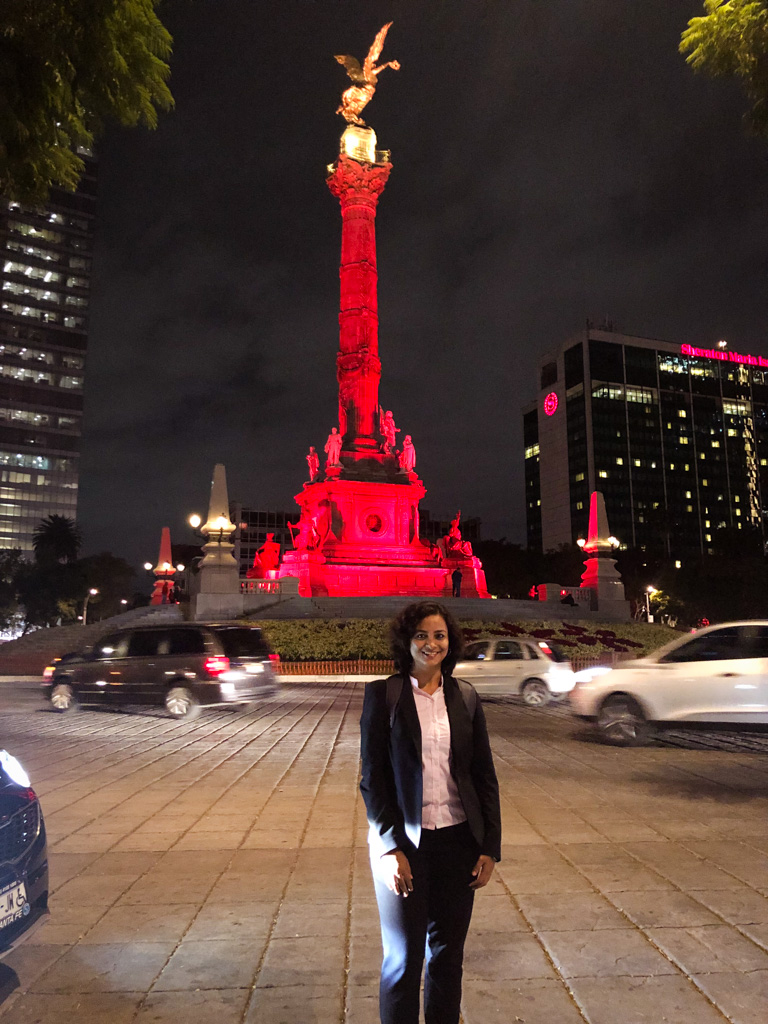 Woman standing in front of the Angel of Independence in Mexico City in the evening, as traffic zoom past in the background.