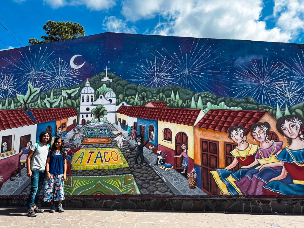 A couple standing with the famous Ataco mural wall in the background. Ruta de las Flores, a must on any El Salvador itinerary.