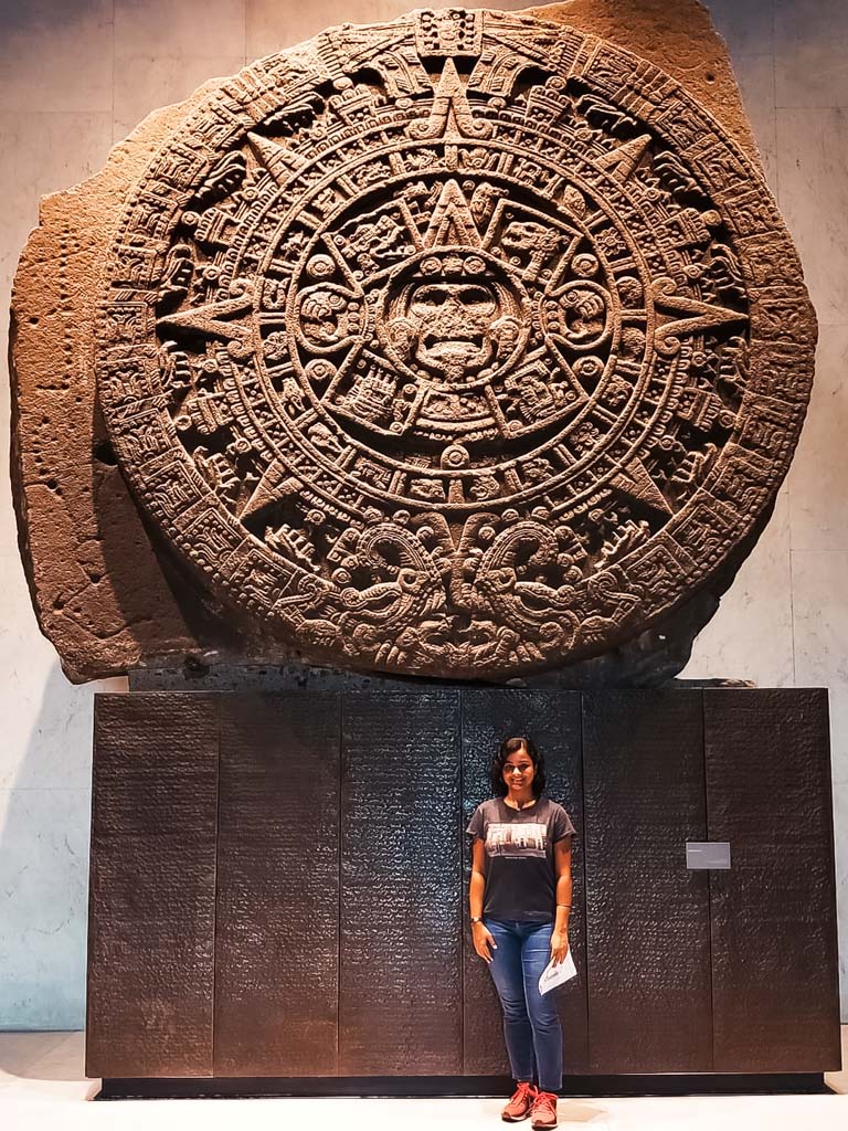 A woman in blue jeans and black grey t-shirt, standing in front of the Sun Stone at the Museum of Anthropology in Mexico City.