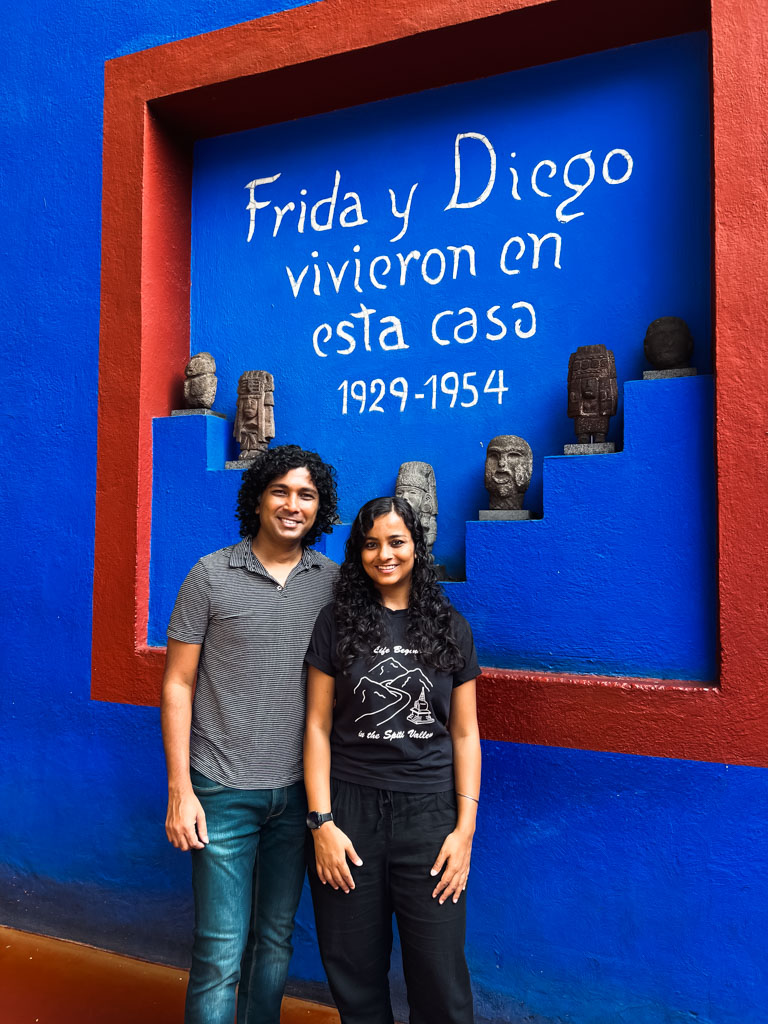 A man and a woman standing next against a blue wall at Casa Azul, where it is written that Frida Kahlo and Diego Rivera used to live here.