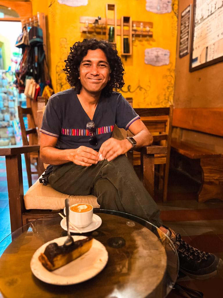 A man at a coffee shop with a cup of coffee and a piece of cake on the table in front of him.