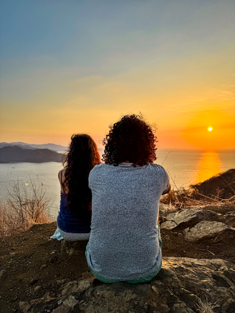 A man and a woman sitting on a cliff watching sunset and the Pacific Ocean near Playa del Coco in Costa Rica. Digital Nomad Visas for Central America and Mexico.