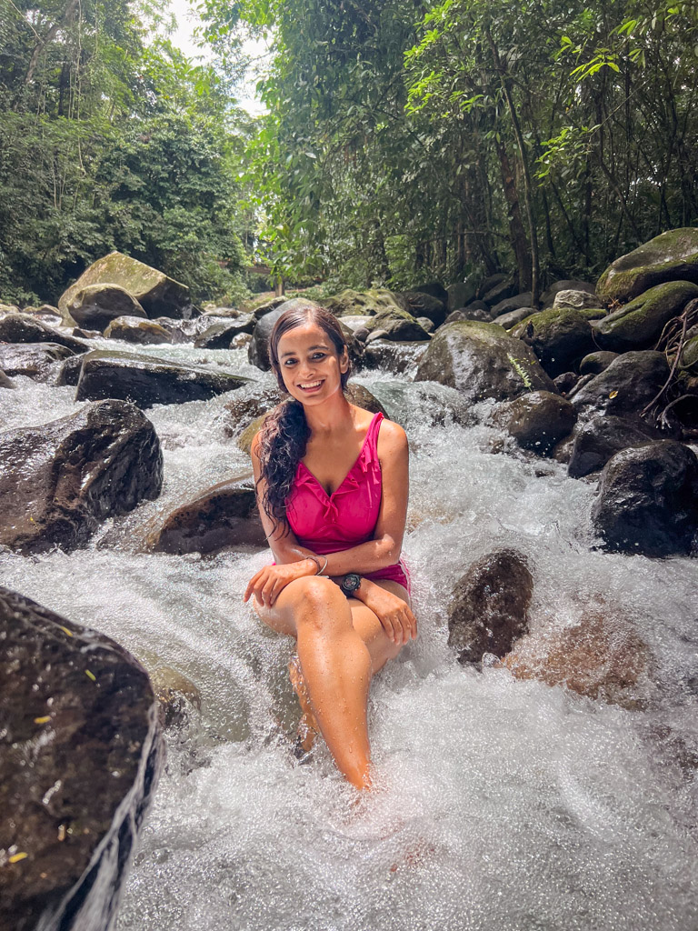 A woman sitting on a large rock in Fortuna river near La Fortuna in Costa Rica. The river water flowing around her, and it is in a forest setting with lots of trees and more rocks in the background. Free Things to Do in La Fortuna.