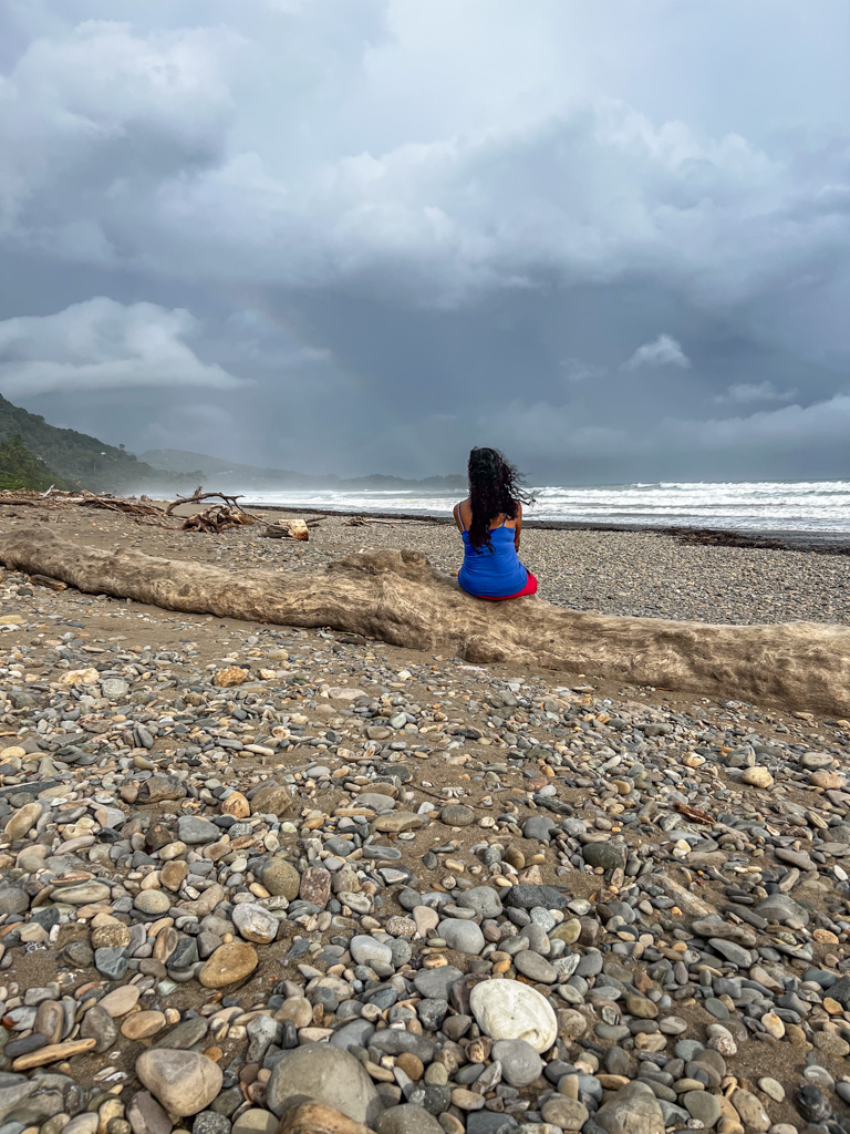 A woman sitting on a dead branch on the rocky Dominical Beach in Costa Rica on a cloudy day. Practicing mindfulness is one of our top well-being tips for Digital Nomads in Latin America. 