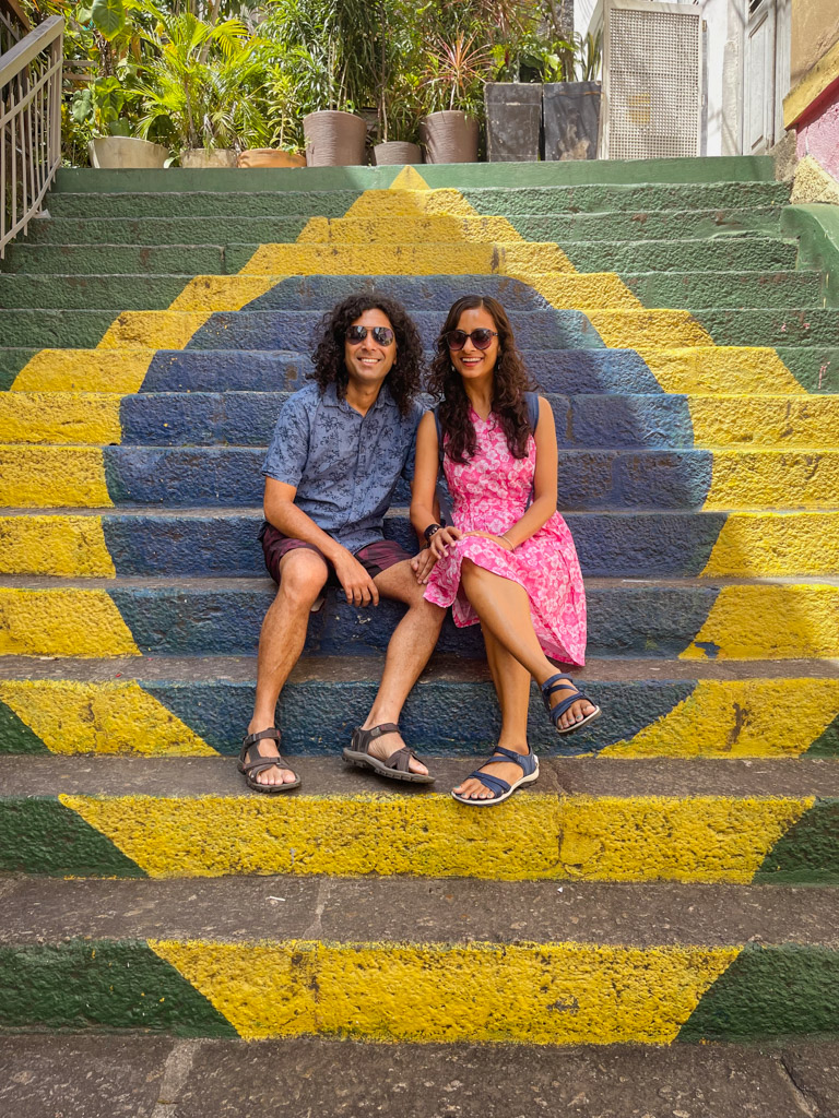 5 Days in Rio de Janeiro Itinerary. A man and a woman seated on a staircase with Brazil flag wall paint.