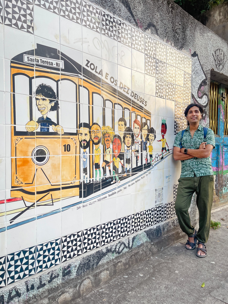 Man standing next to an interesting wall art in Santa Teresa. Wall art representing classic Santa Teresa tram with the world's best footballers on board.
