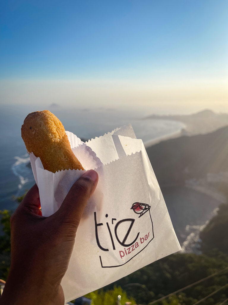 Small bites with big views, a hand holding a snack with view of Rio de Janeiro from Sugarloaf mountain.