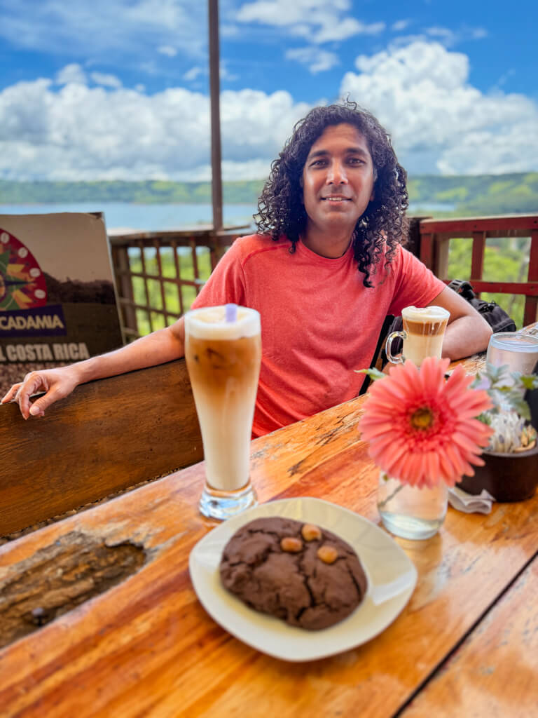 A man sitting in Cafe Macadamia with a cold coffee and a chocolate cookie on the table in front of him during a stop while driving between La Fortuna to Monteverde.