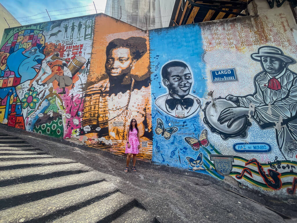 A woman in pink floral dress standing next to an important wall mural at Pedra Do Sal, the birthplace of Samba music in Rio de Janeiro.