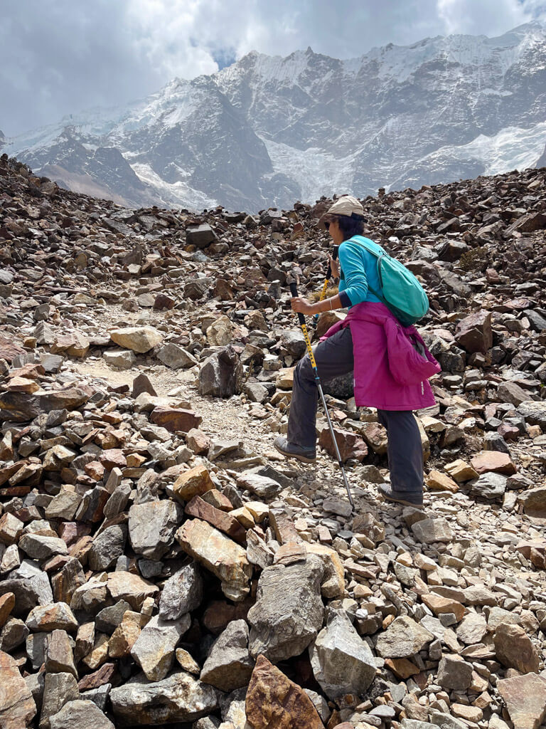 A woman walking up a rocky mountain path. Snow capped mountain in the backdrop. Dressing in layers is an important strategy when packing for Salkantay Trek.