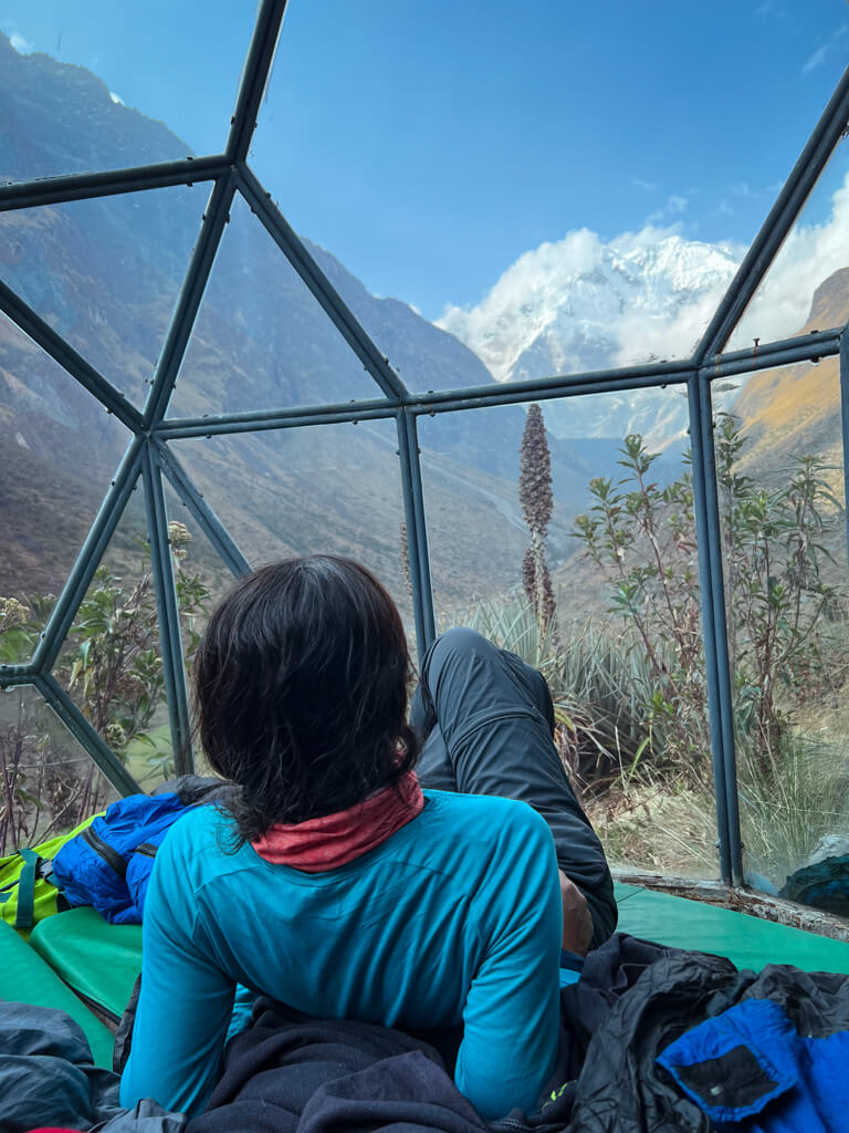 A woman half lying at a Salkantay Trek campsite and admiring the view of a snowcapped mountain.