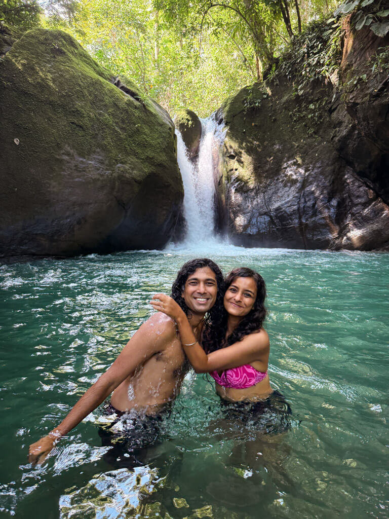 A man and a woman standing in a river with the Cascada El Pavon waterfall behind, near Uvita, Costa Rica.