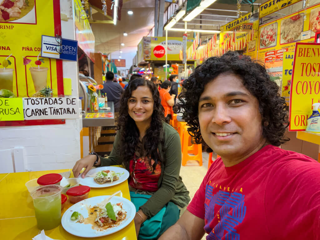 A man and a woman, with plates of tostadas on their table, at one one of the stalls in Coyoacan Market.
