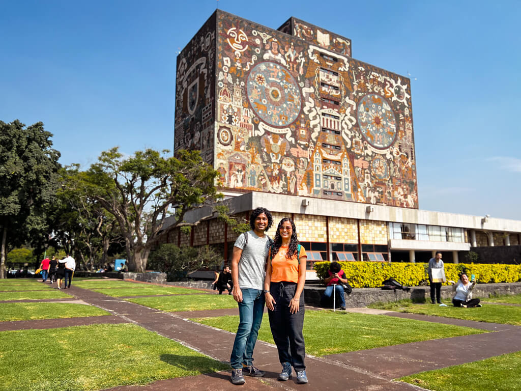 A man and a woman standing in the campus of UNAM. Behind them is a building with giant mural work. They are wearing comfortable pants, tees and walking shoes, essentials on the list for what to pack for Mexico City.