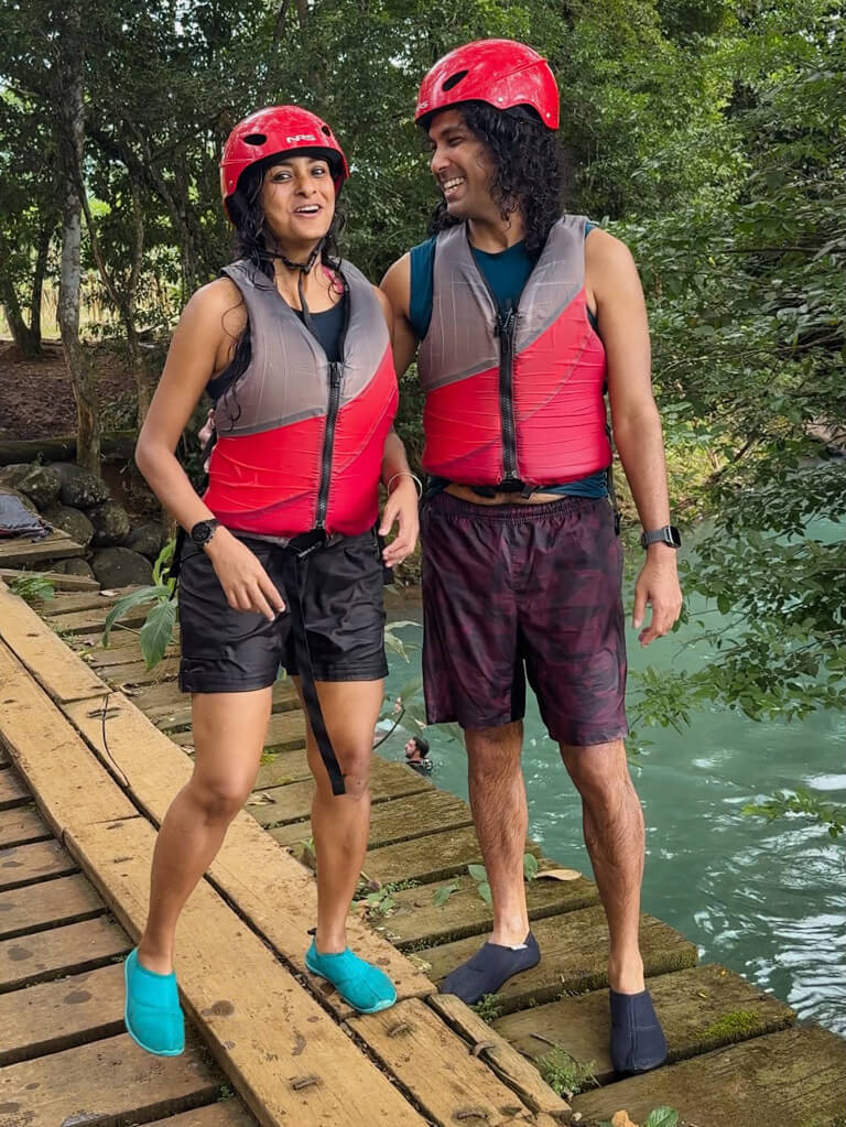A man and a woman wearing wet clothes, water shoes, life jackets and helmets, standing on a wooden bridge over the river, Rio Celeste in Costa Rica.
