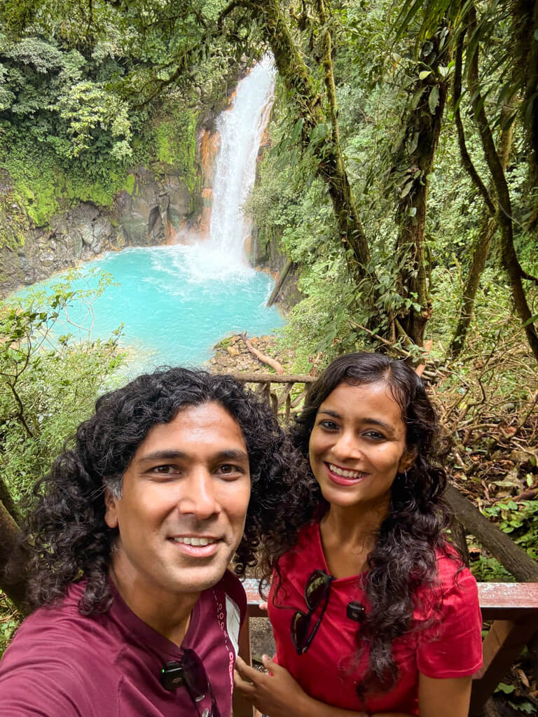 A woman and a man standing on the stairs that lead to the Rio Celeste Waterfall, visible behind them. The water of the pool is bright blue colored and surrounded by a forest.