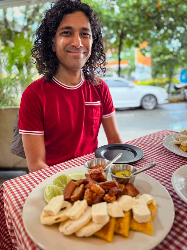 A man in red t-shirt seated at a table with red and white checkered table cloth. On the table, there is a plate with chicharron, chorizo, arepa blanca, arepa de chocolo, guacamole, hogao, all part of the tastings on a Local Food Tour in Medellin.
