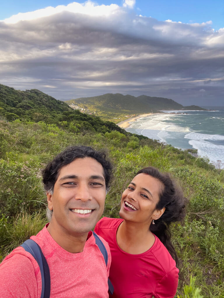 A man and woman on a hiking trail on a hill with the view of a beach and ocean in the background, in Florianopolis.