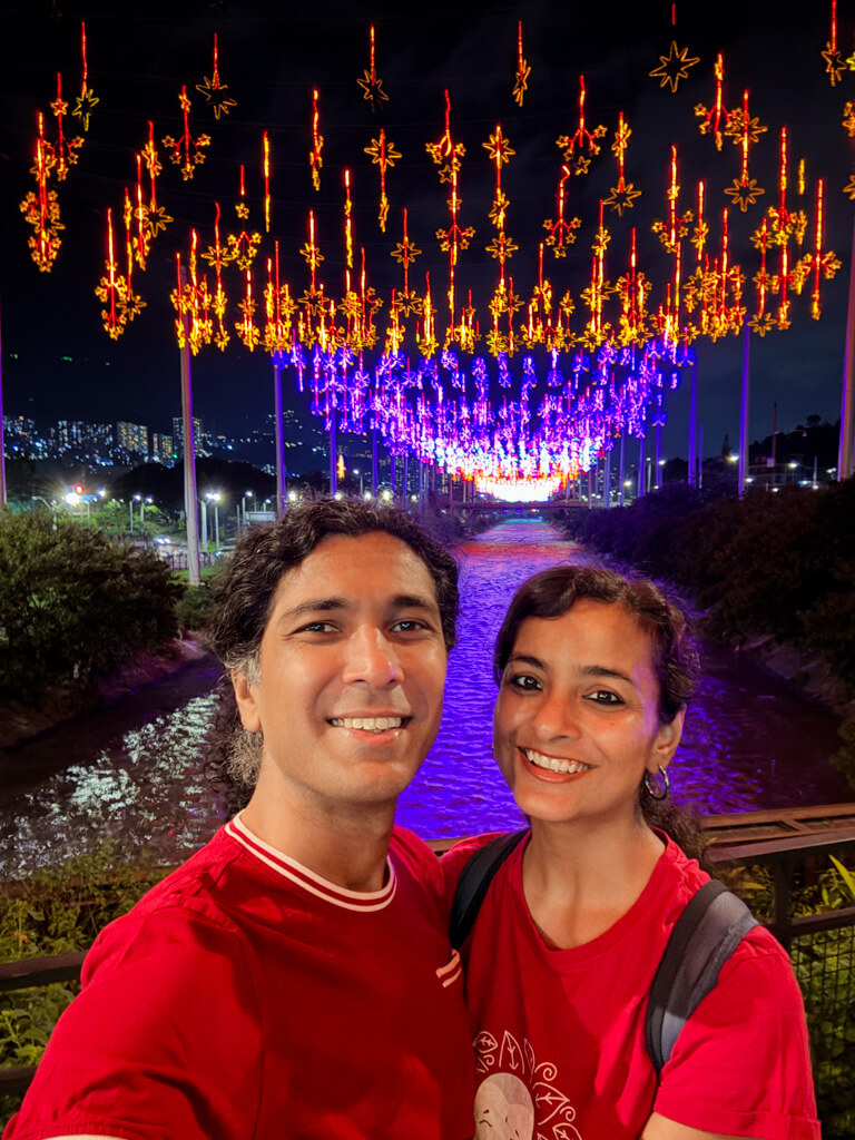 A man and a woman, standing on the bridge at Parques del Rio in Medellin. Christmas lights decoration in the background.