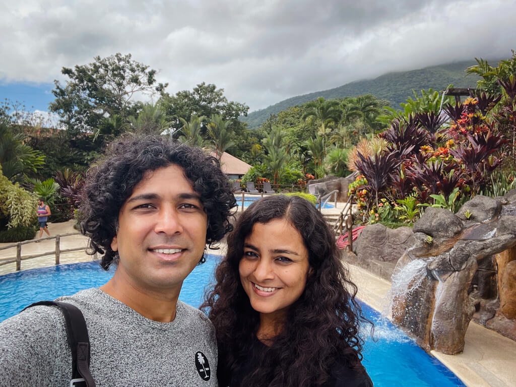 A man and a woman, standing in front of a pool at Los Lagos Hotel. Lush landscaping and nature surrounding the pool area.