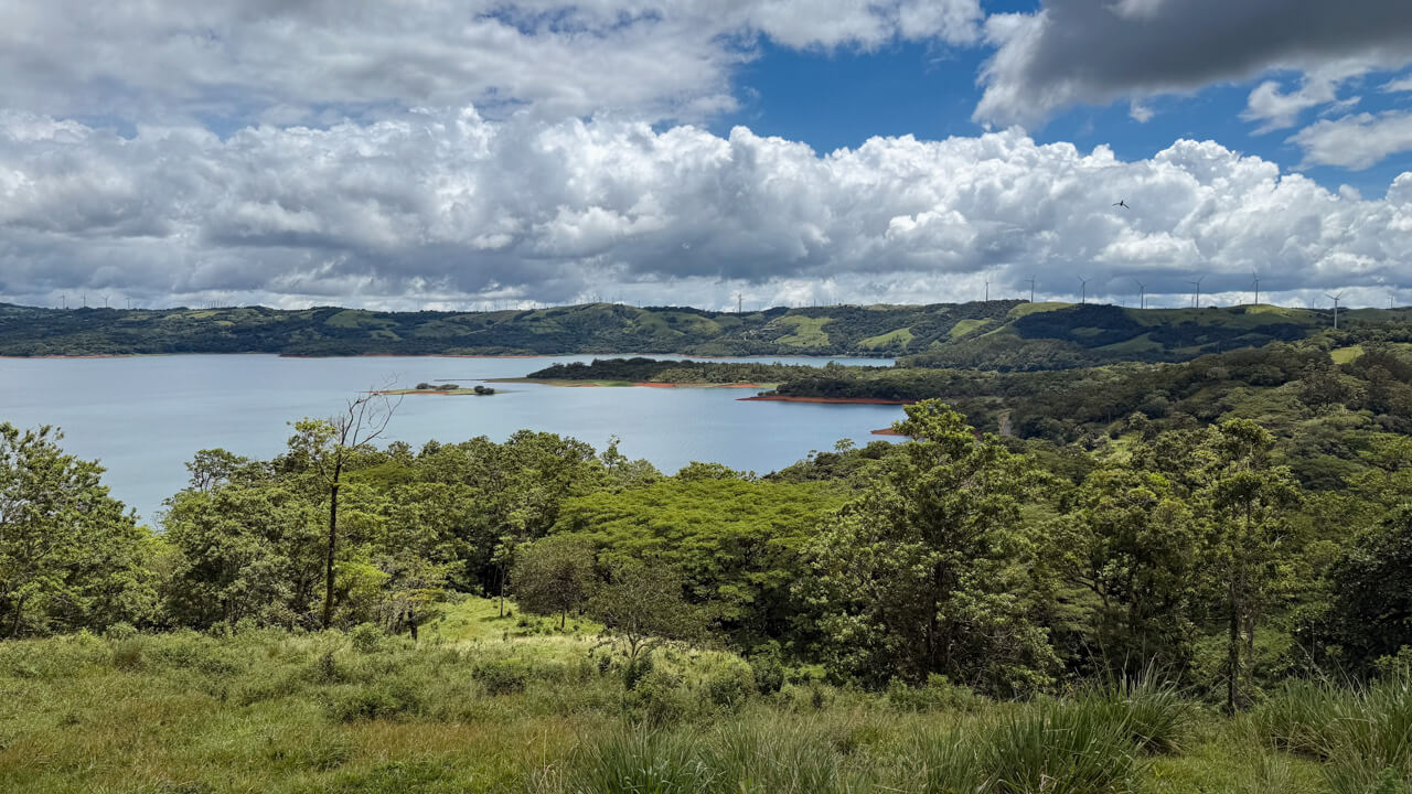 A scenic view of green foliage, mountains, lake, windmills and blue sky. at a pitstop when driving from La Fortuna to Monteverde.