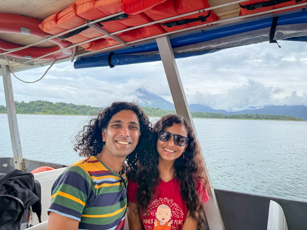 A man and woman in a boat ride on the way from La Fortuna to Monteverde across Arenal Lake with Arenal Volcano partially visible behind, between clouds.