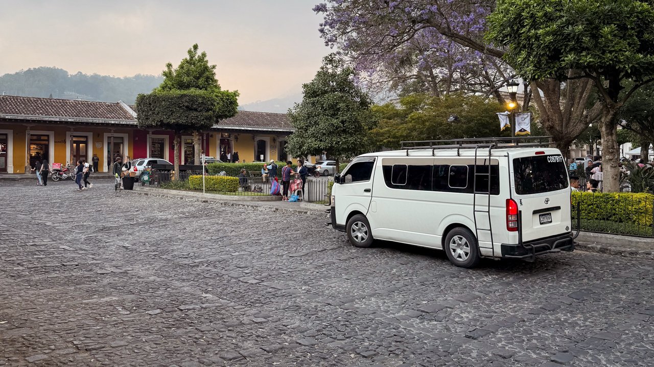 A white shuttle minivan parked on the road next to Parque Central in Antigua Guatemala.
