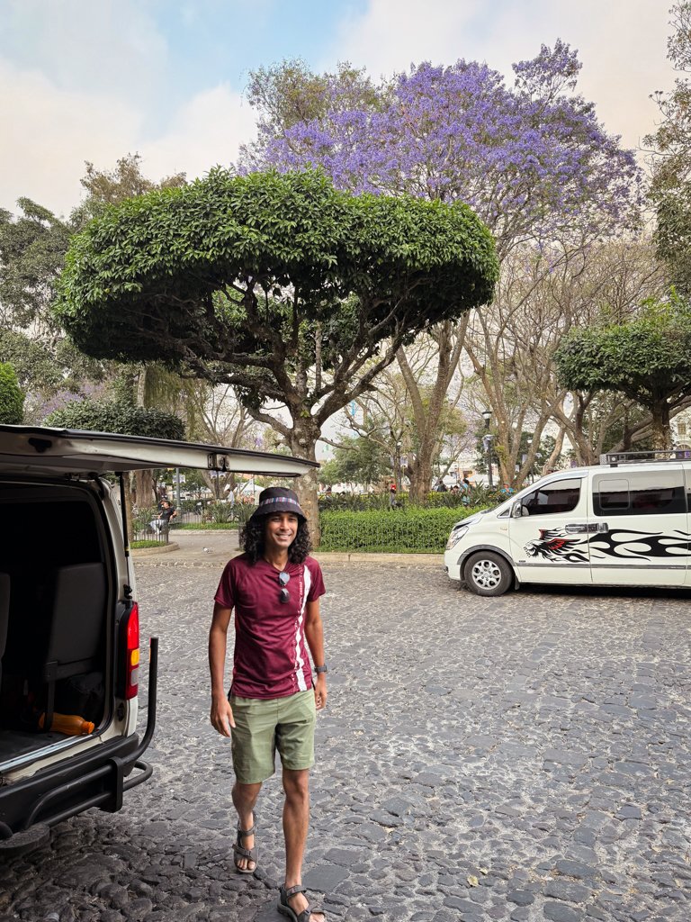 A man in a maroon quick-dry t-shirt and green shorts, and wearing hiking sandals, walking next to a shuttle minivan in Antigua Guatemala, after arriving from El Paredon.